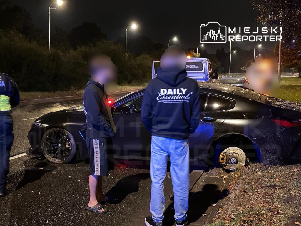 Mężczyzna w dresie i klapkach rozmawia z policjantem obok czarnego samochodu z uszkodzoną oponą na nocnej ulicy.