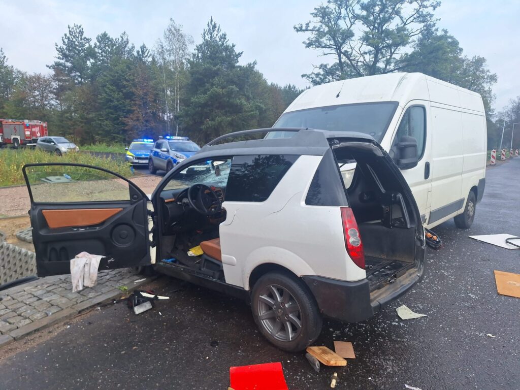 W wyniku wypadku, uszkodzony samochód osobowy stoi na drodze, obok białego furgonu; w tle widoczne pojazdy policyjne.
