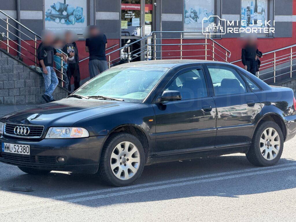 Czarny samochód osobowy marki Audi zaparkowany na ulicy, obok grupka ludzi rozmawiających na schodach budynku.