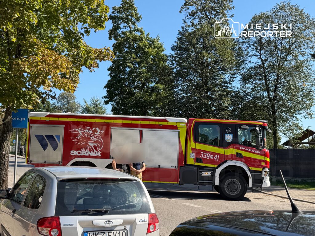 Wóz strażacki marki Scania zaparkowany przy ulicy, obok policjantów rozmawiających z przechodniami.