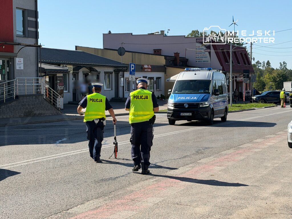 Dwaj policjanci w odblaskowych kamizelkach idą ulicą obok radiowozu policyjnego w otoczeniu budynków handlowych.