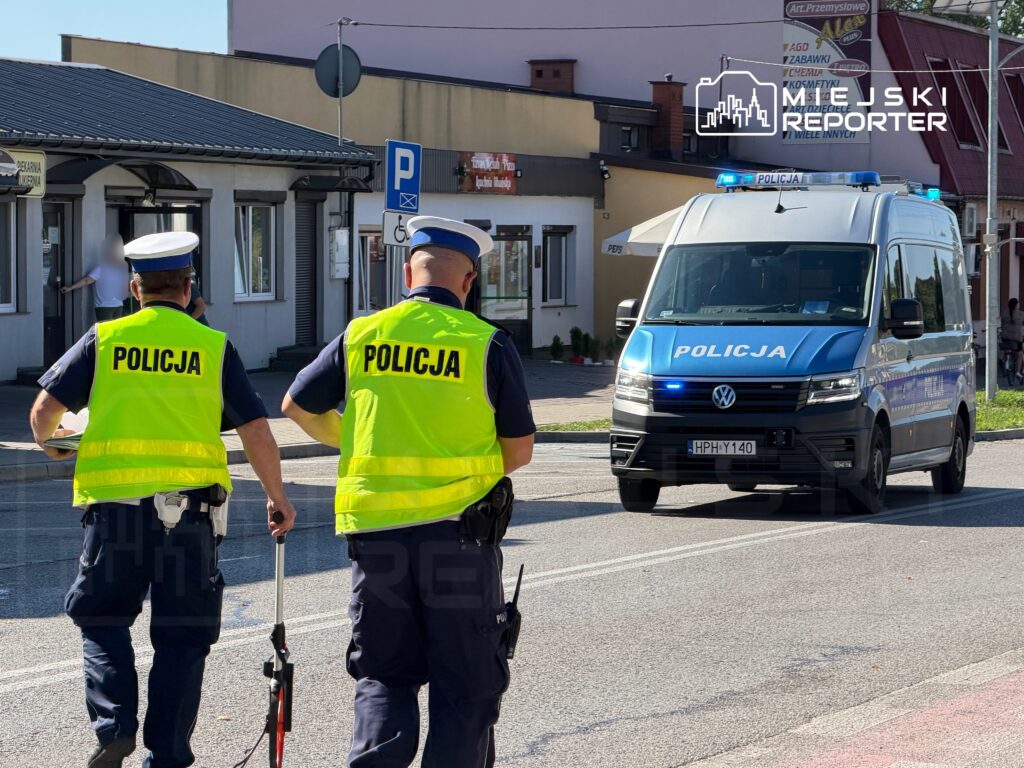 Dwóch policjantów w odblaskowych kamizelkach kontroluje ruch na ulicy, obok przejeżdża radiowóz policyjny.