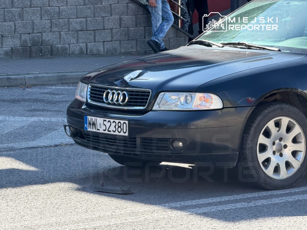 Czarny samochód marki Audi zaparkowany na ulicy, z widoczną tablicą rejestracyjną. W tle przechodnie na schodach.