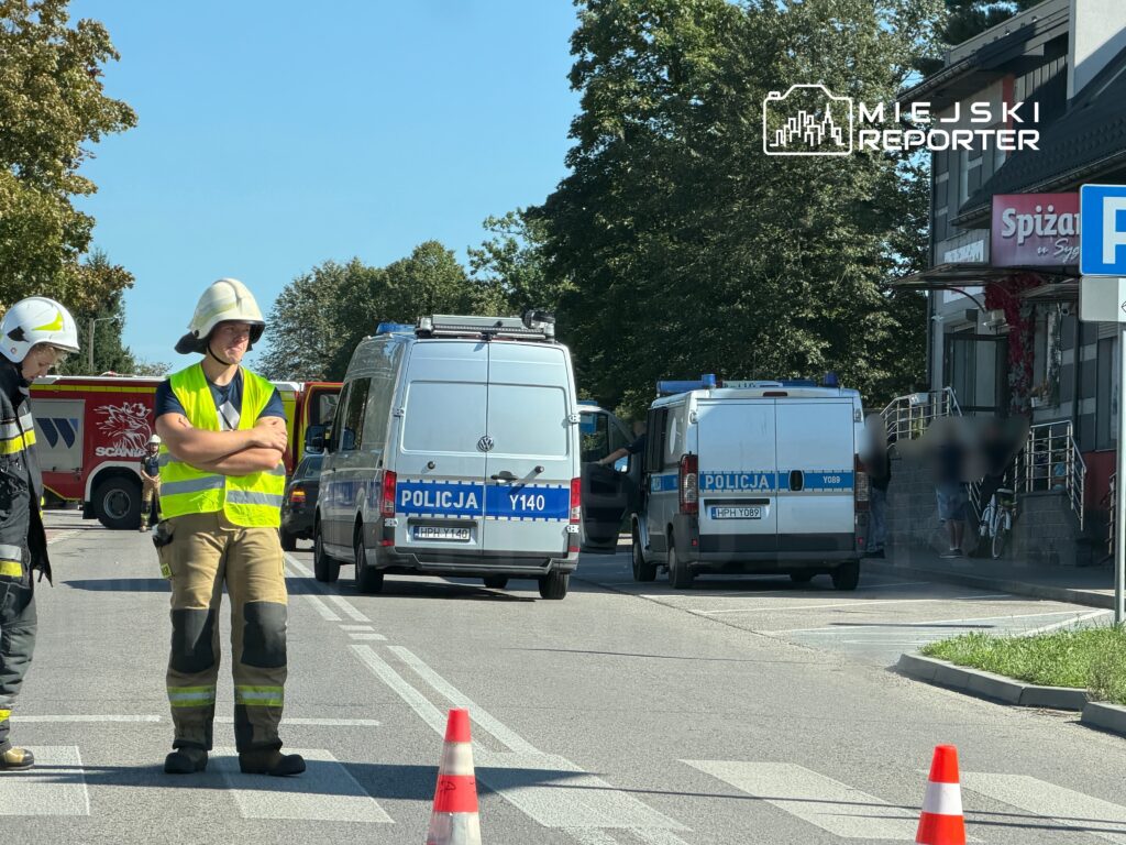 Strażak w hełmie i kamizelce odblaskowej obserwuje akcję ratunkową przy budynku z czerwonymi kwiatami.