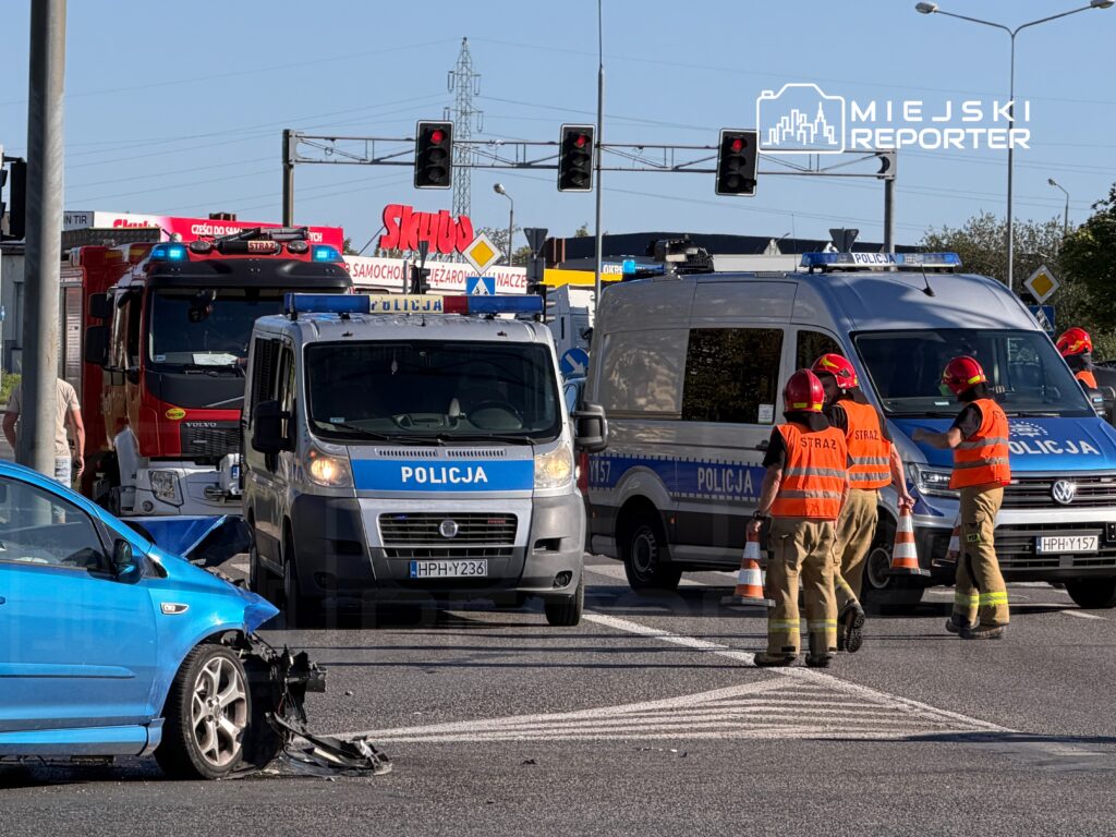 W wyniku wypadku drogowego na skrzyżowaniu, niebieski samochód z uszkodzoną przednią częścią stoi obok pojazdów służb ratunkowych.