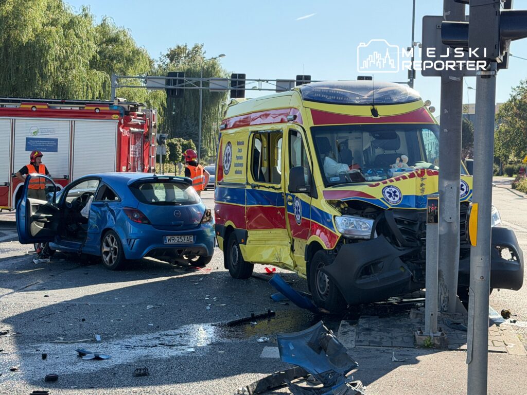 Na skrzyżowaniu leży uszkodzony niebieski samochód osobowy oraz ambulans, obok pracują strażacy w pomarańczowych kamizelkach.