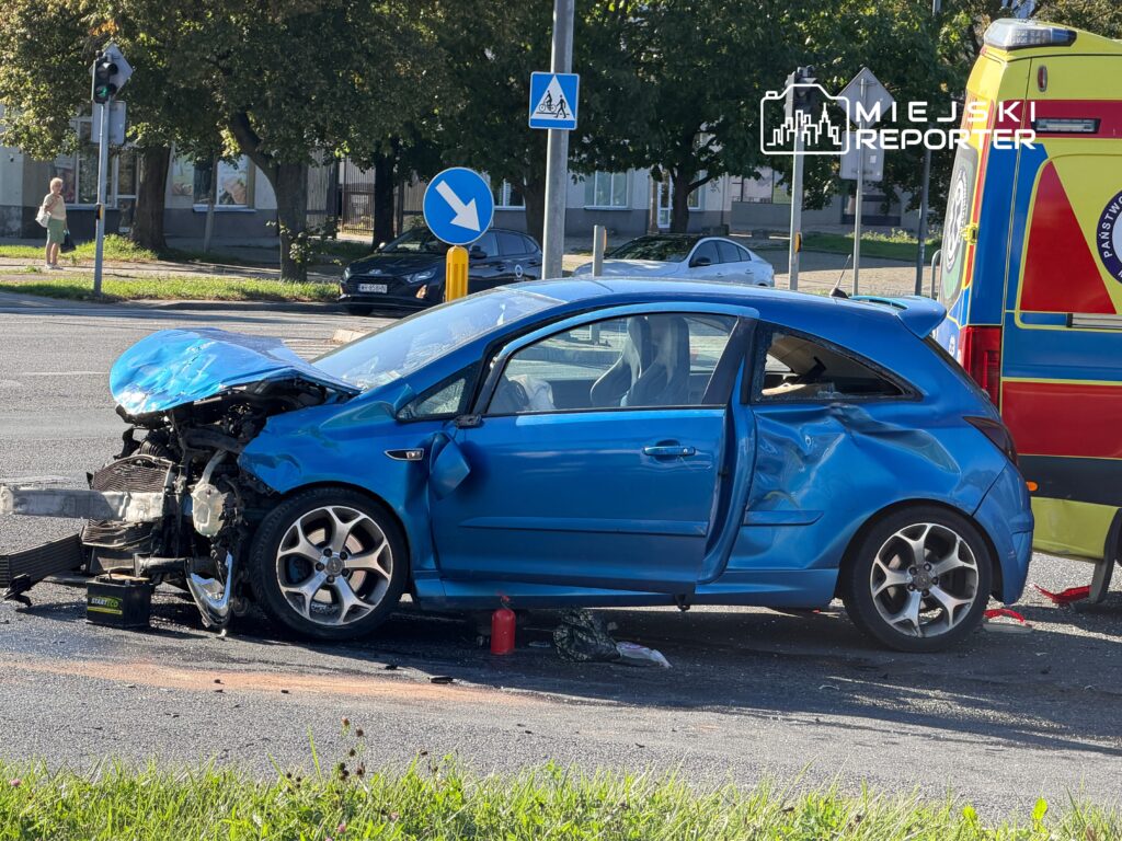 Niebieski samochód osobowy z uszkodzoną przednią częścią stoi na drodze, obok karetka pogotowia.