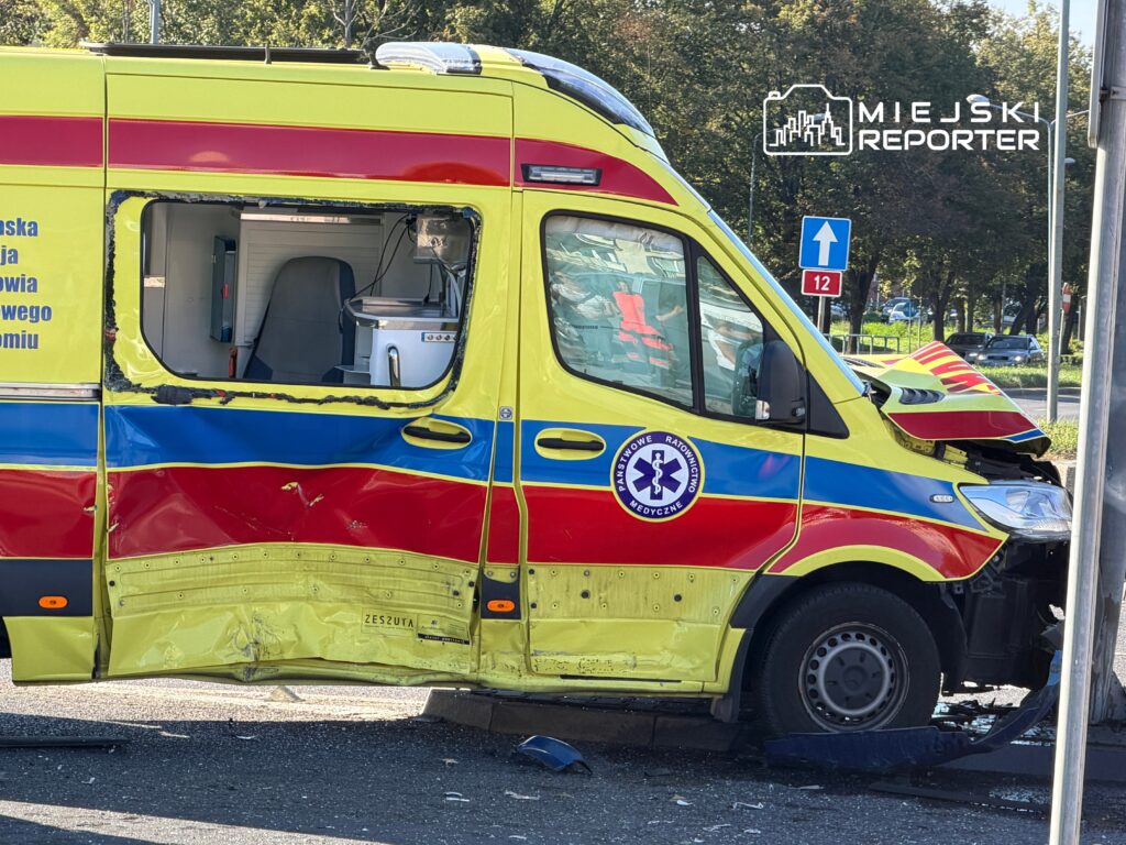 Uszkodzony ambulans z wgniecioną boczną częścią stoi na ulicy, obok znaku drogowym.