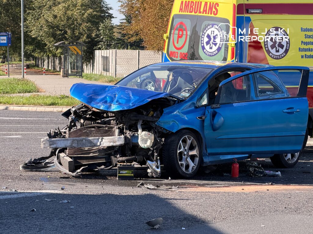 Uszkodzony niebieski samochód osobowy stoi na drodze, obok niego widać ambulans oraz przystanek autobusowy.