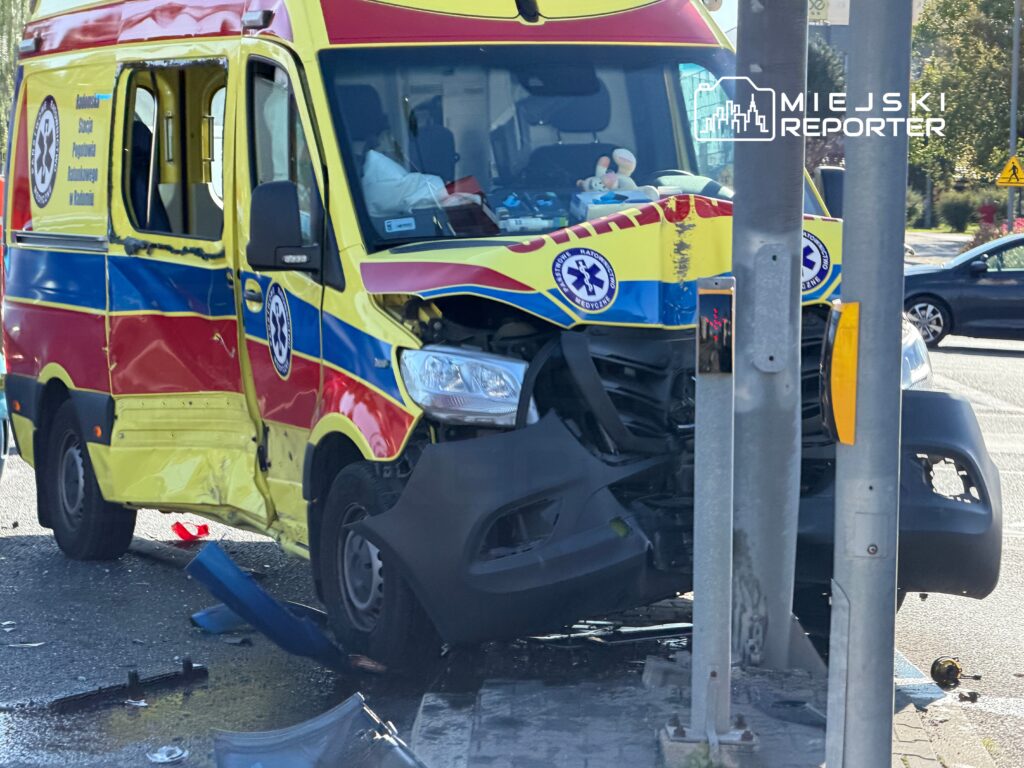 Karetka pogotowia z uszkodzoną przednią częścią stoi przy latarni ulicznej na skrzyżowaniu. Wokół porozrzucane fragmenty pojazdu.