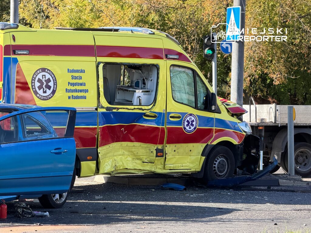 Zderzenie karetki pogotowia z niebieskim samochodem osobowym na ulicy, uszkodzona strona karetki widoczna z bliska.