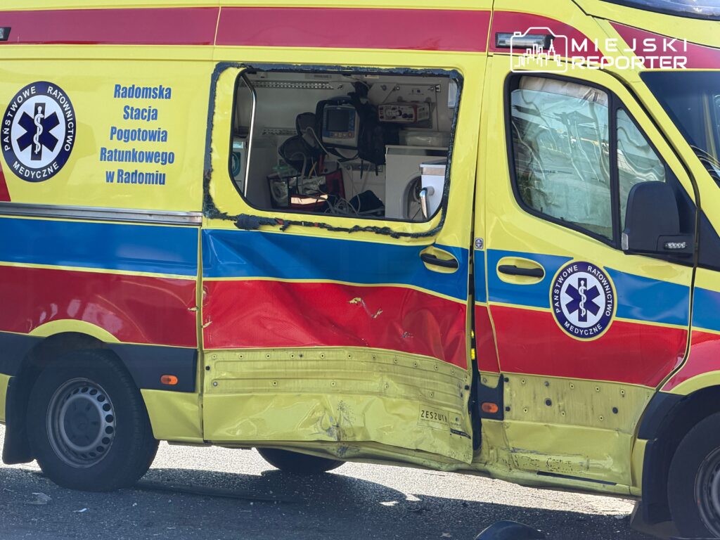 Uszkodzona karetka pogotowia z widocznymi śladami zderzenia, zaparkowana na ulicy. Na drzwiach widnieje napis "Radomska Stacja Pogotowia Ratunkowego w Radomiu".