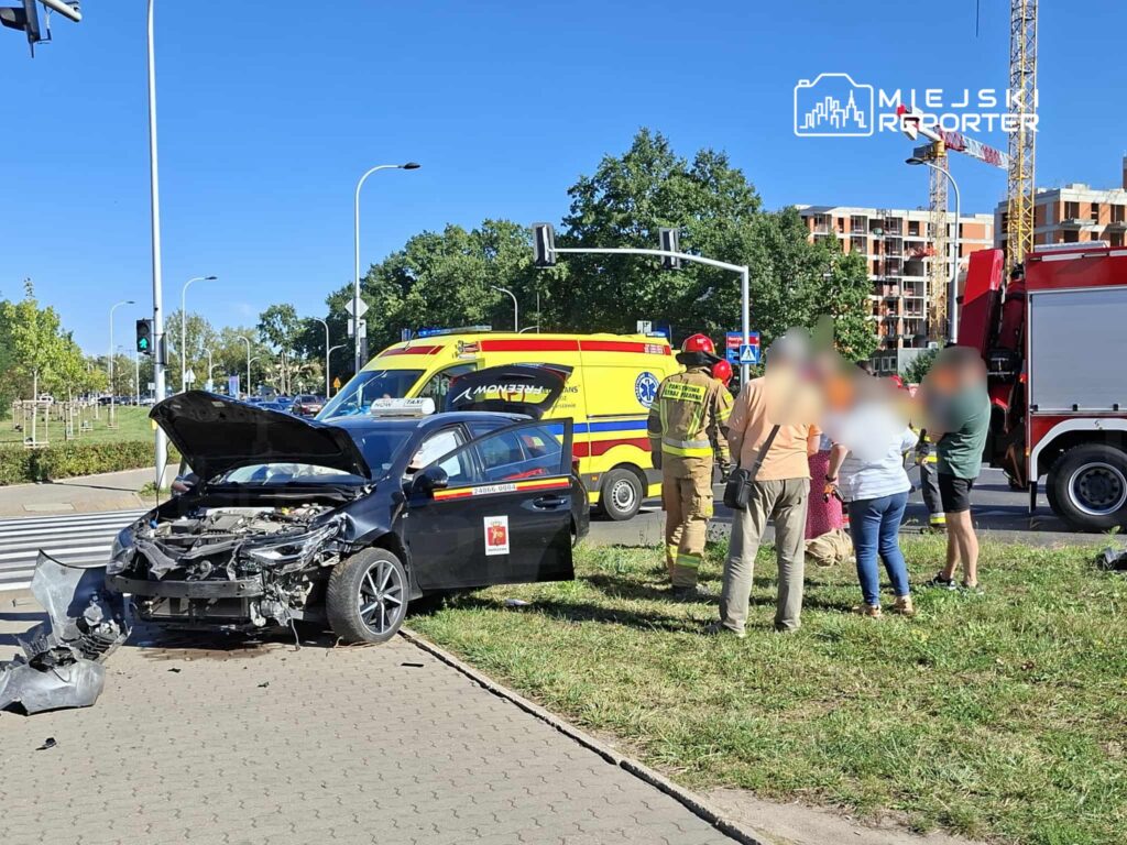 Czarny samochód osobowy z uszkodzoną maską stoi na trawie obok jezdni, wokół niego grupa osób oraz służby ratunkowe.