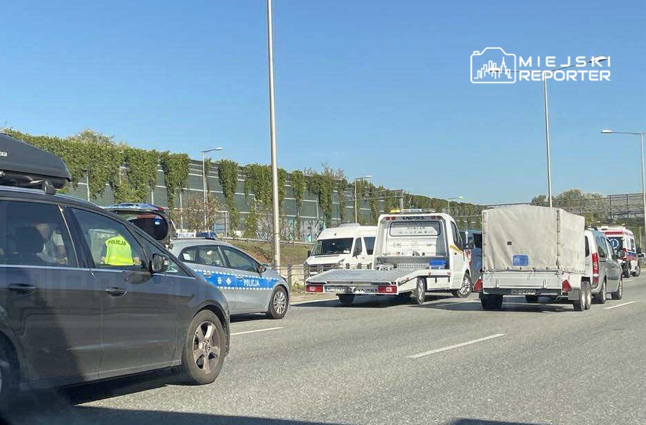 Ruch drogowy zablokowany przez kilka pojazdów, w tym radiowozy policji oraz samochody dostawcze, na szerokiej jezdni.