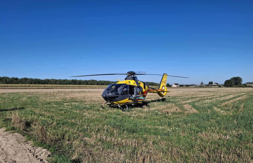 Śmigłowiec ratunkowy lądujący na polu w otoczeniu zielonych łąk i drzew, pod jasnym niebem.