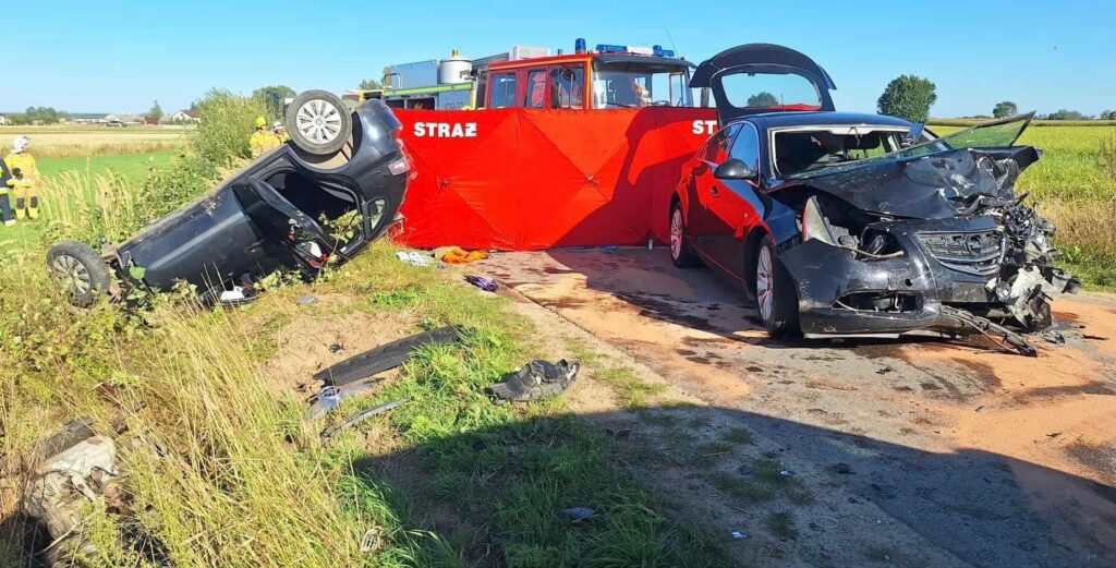 Wypadek drogowy z przewróconym samochodem osobowym oraz rozbitym autem na poboczu drogi, w tle straż pożarna.