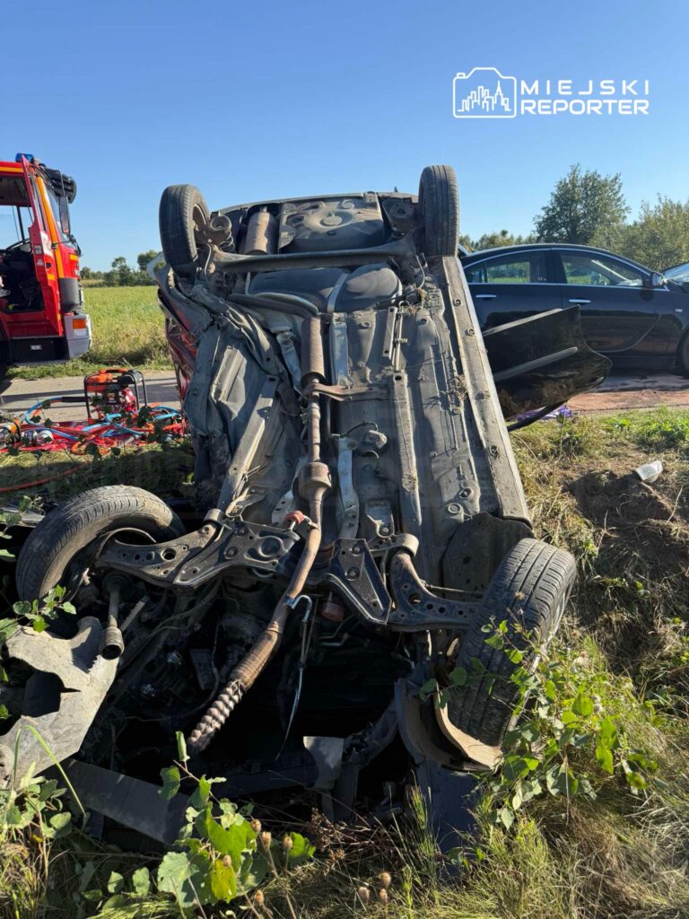Odwrócone auto osobowe leży na poboczu drogi, otoczone trawą i krzewami, obok widać wóz strażacki.