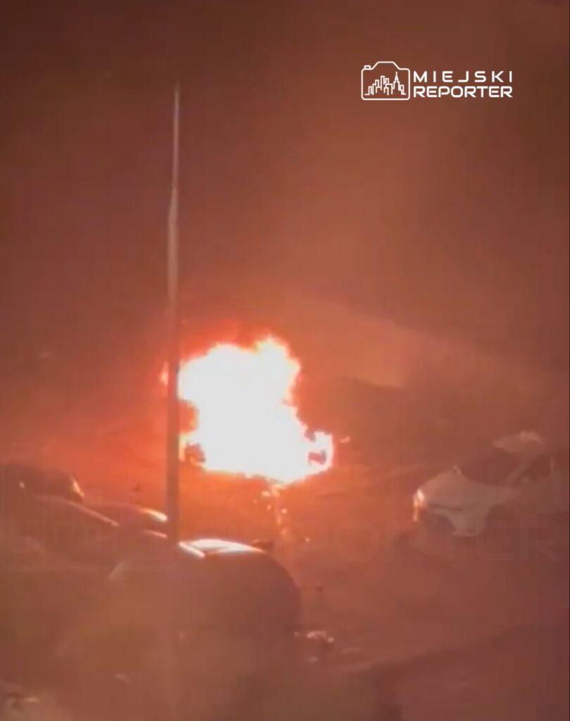 Pojazd płonący na parkingu, otoczony przez inne samochody w ciemnej scenerii nocnej.