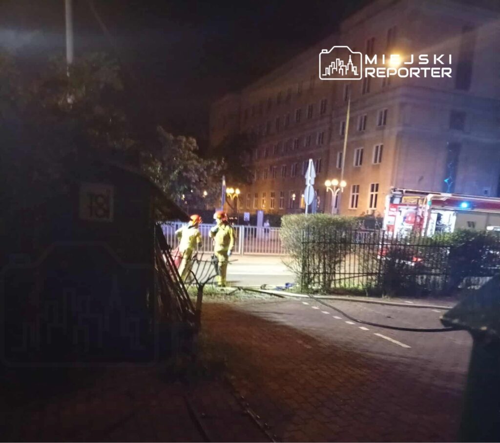 Dwaj strażacy w żółtych mundurach pracują nocą przy włączonym wozie strażackim w mieście.