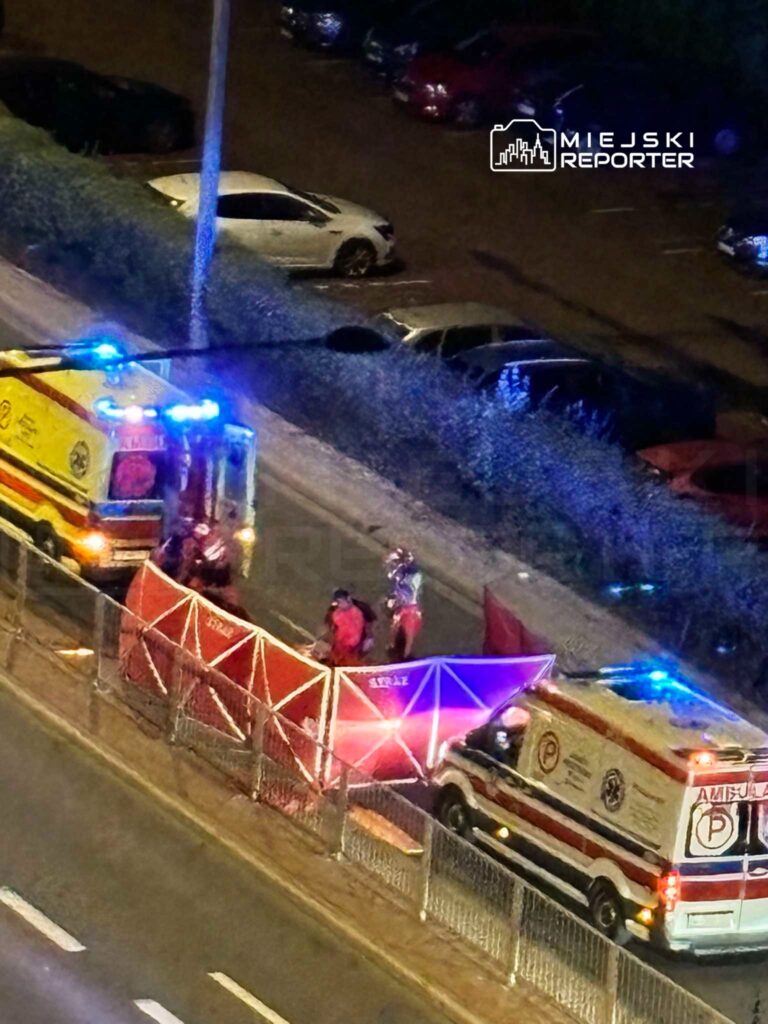 Zespół ratunkowy ustawia namiot medyczny obok ambulansów na ulicy w nocy, otoczeni przez samochody zaparkowane w tle.