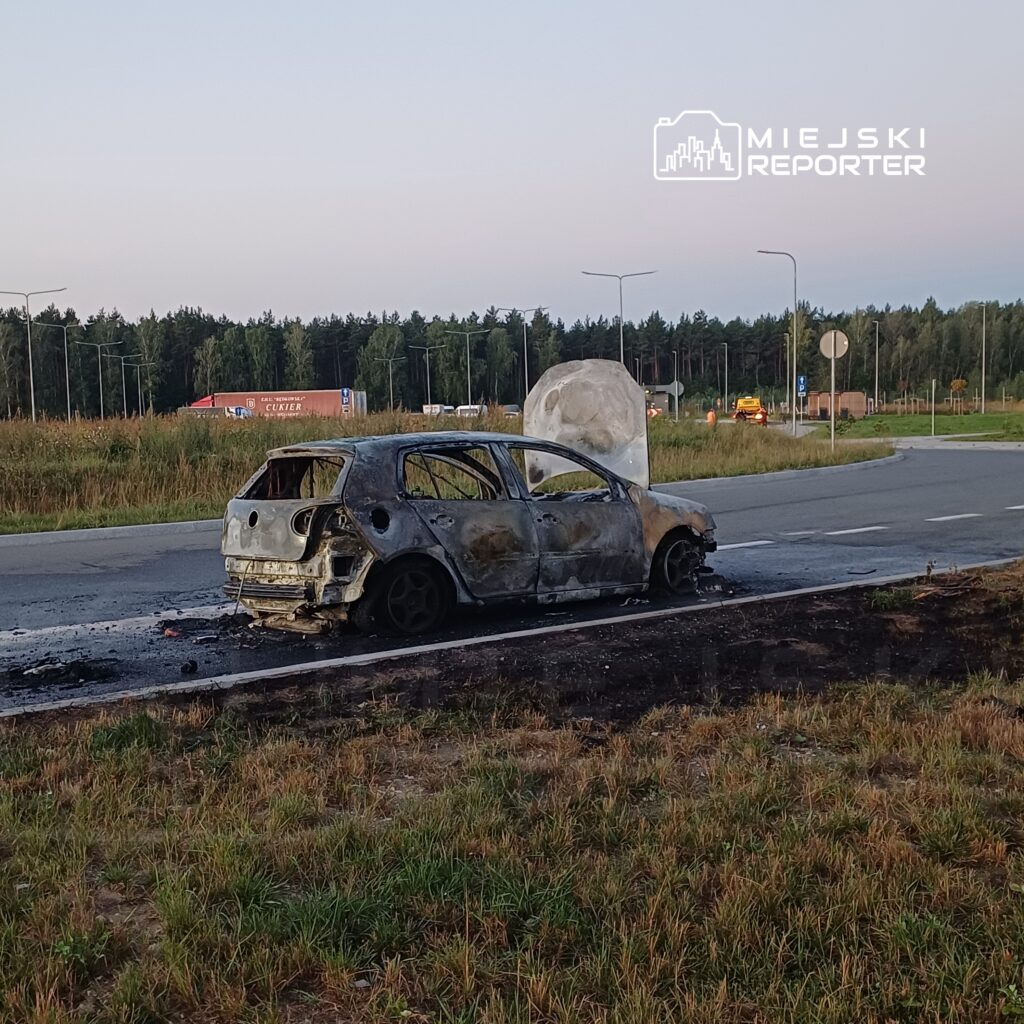Spalony samochód osobowy zaparkowany na poboczu drogi, obok widoczna zieleń i znaki drogowe.
