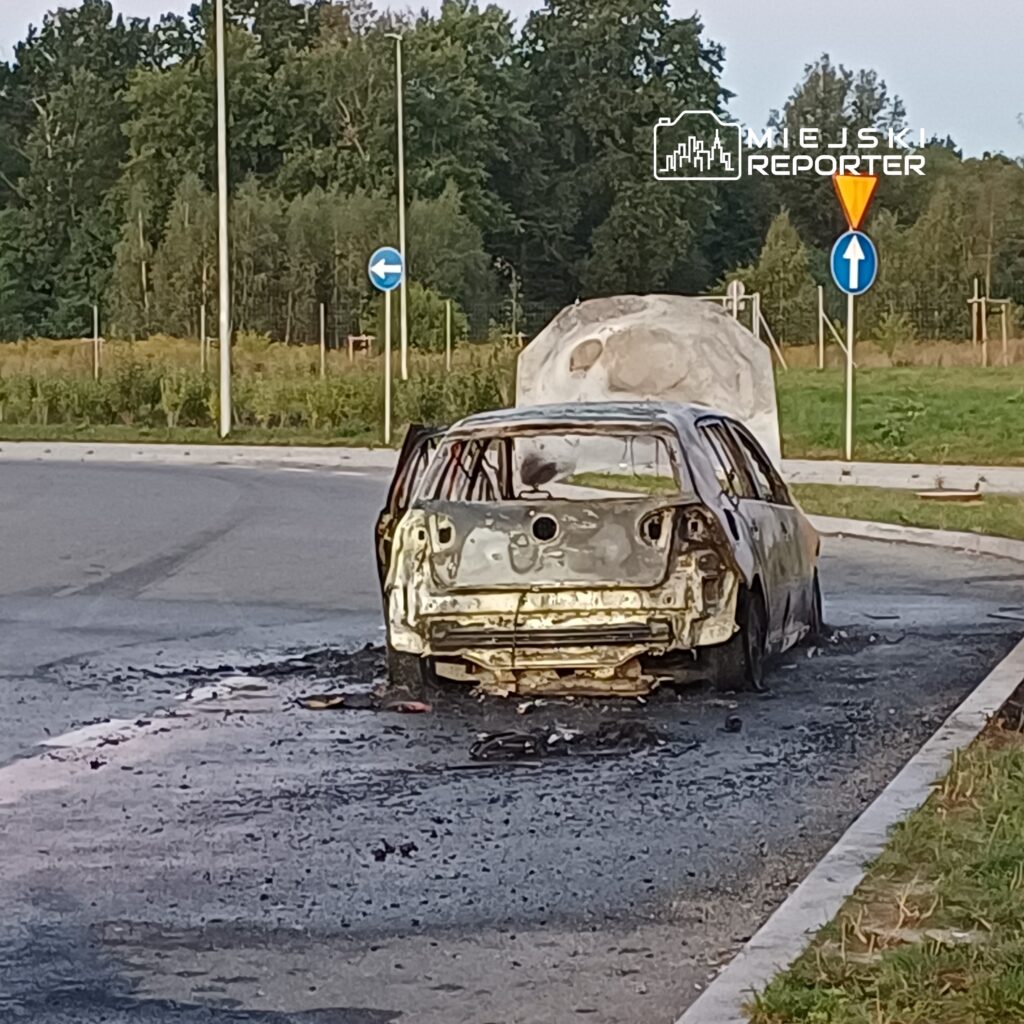 Spalony samochód osobowy stoi na poboczu drogi w otoczeniu zieleni i znaków drogowych.
