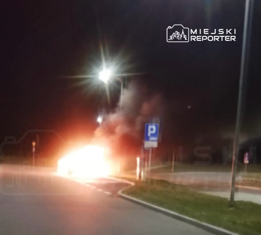 Pojazd płonący na poboczu drogi, otoczony gęstym dymem, w nocy pod oświetleniem ulicznym.