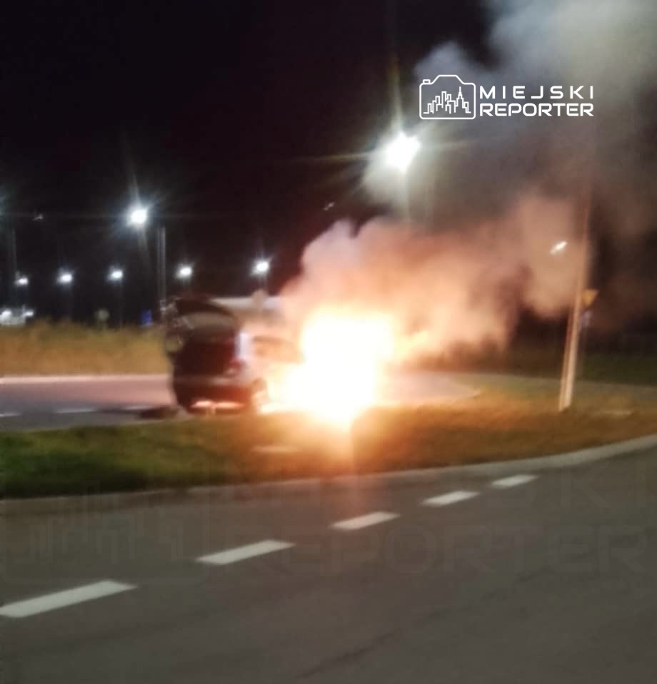 Samochód płonący na rondzie, w tle w nocy widoczne oświetlenie uliczne oraz zarys zieleni.