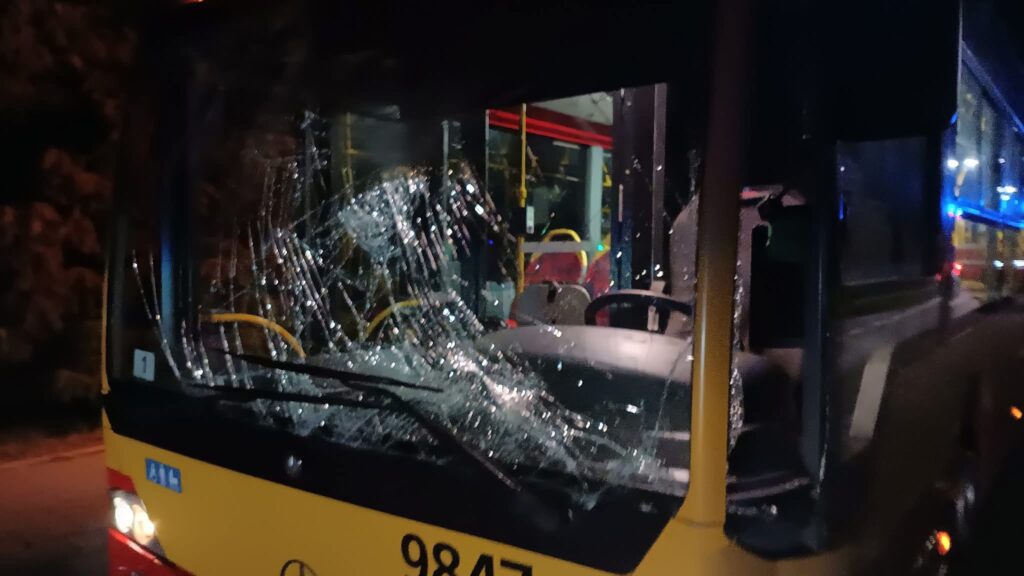 Zniszczona szyba przednia autobusu z widocznymi pęknięciami w nocy, w tle oświetlona przestrzeń przystanku.
