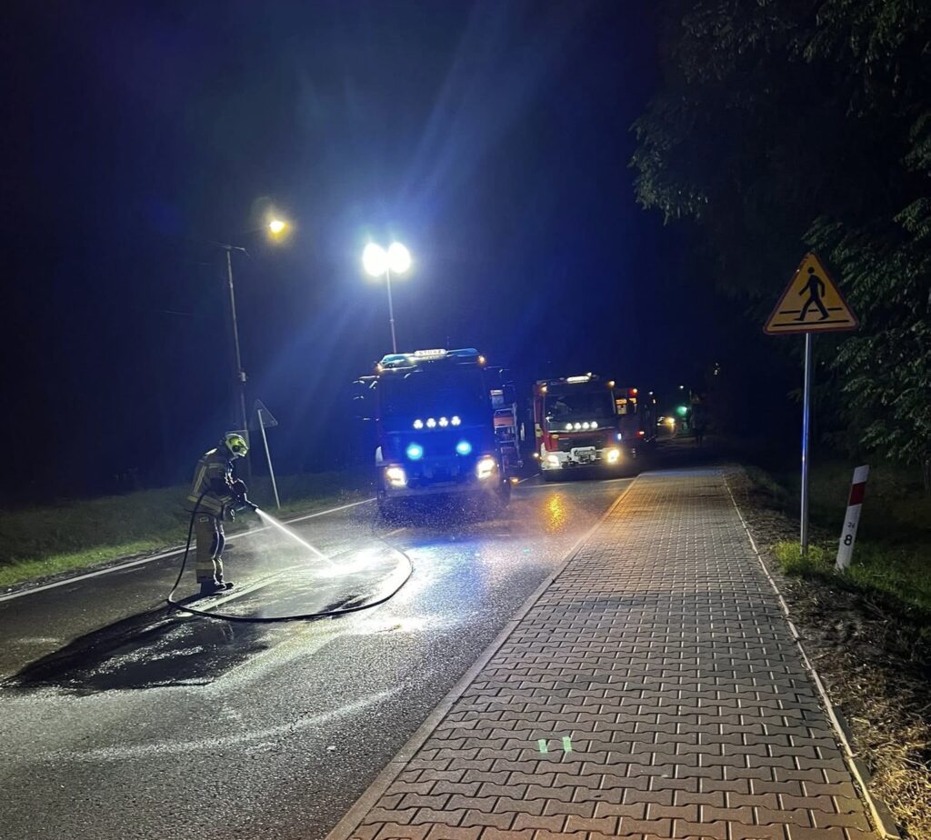 Strażak w hełmie myje asfalt na drodze, obok stoją wozu strażackie z włączonymi sygnałami świetlnymi.