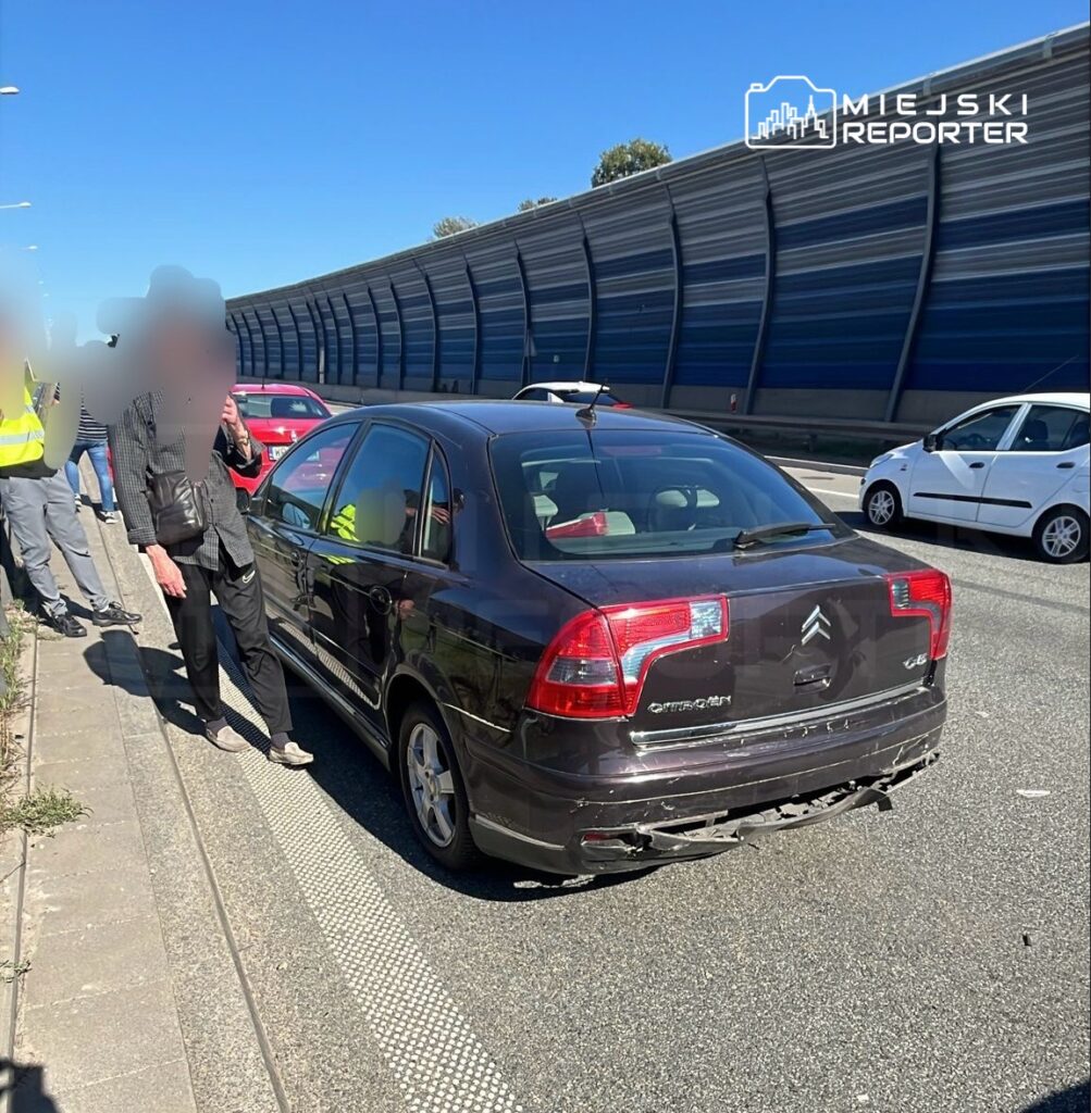 Samochód osobowy marki Citroën zatrzymany na pasie drogowym, obok grupy osób w odzieży roboczej, w tle widoczna droga.