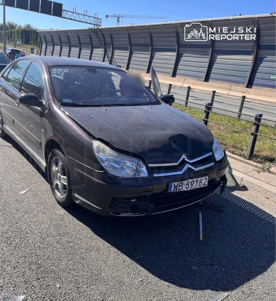 Czarny samochód osobowy z uszkodzoną przednią częścią stoi na poboczu drogi, obok leży narzędzie.