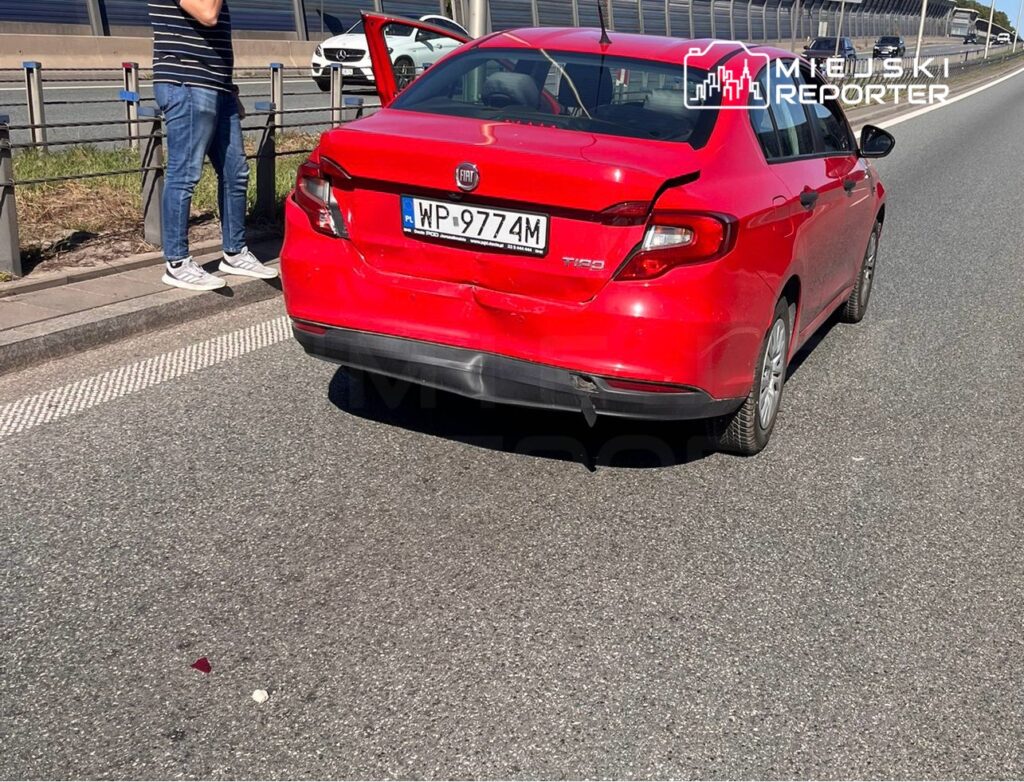 Czerwony samochód osobowy z uszkodzeniami tyłu stoi na poboczu drogi, obok mężczyzna w koszulce w paski rozmawia przez telefon.