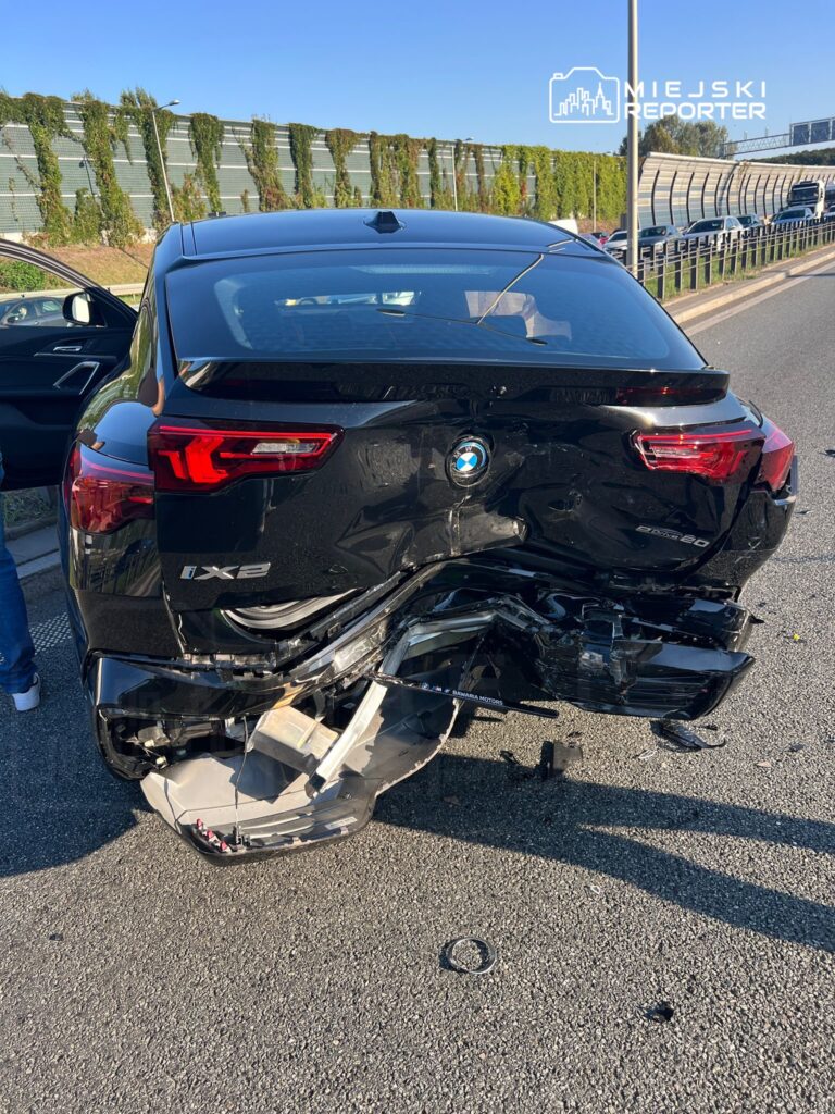 Czarny samochód marki BMW z uszkodzeniem tyłu stoi na drodze, obok niego kilka osób rozmawia.
