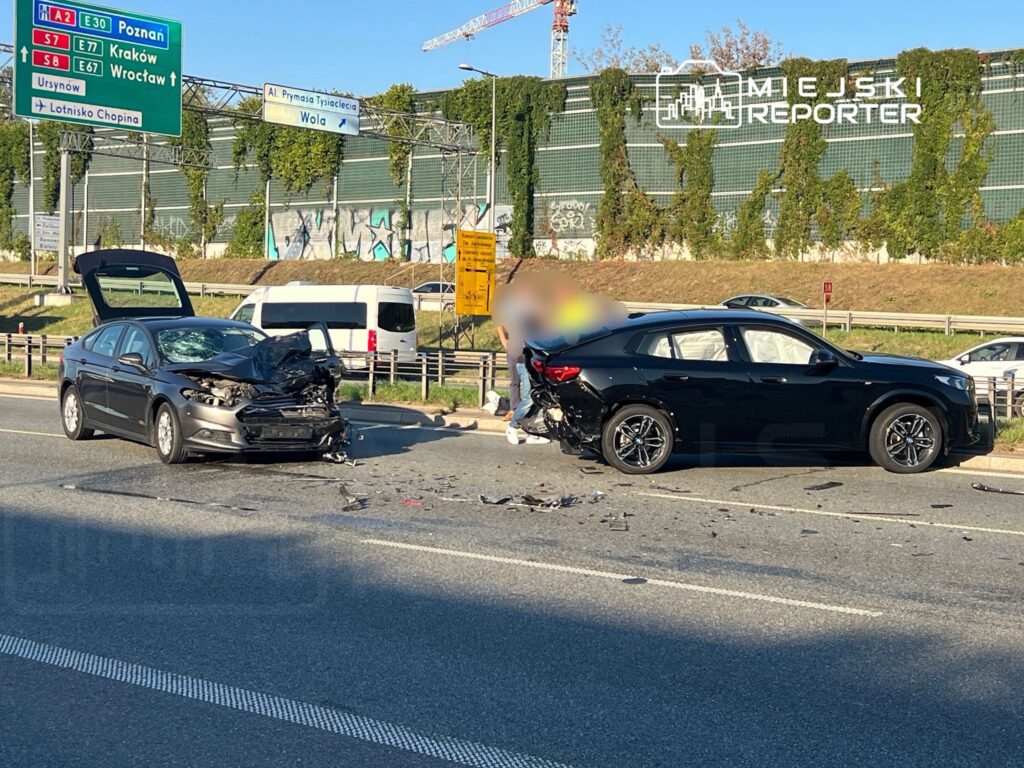Dwa uszkodzone samochody osobowe na ruchliwej drodze, jeden z otwartą maską, w tle znaki drogowe.