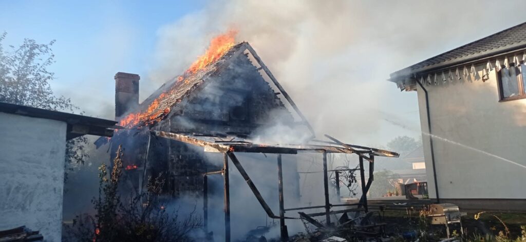 Płonący dach drewnianego budynku, dym i ogień wydobywają się z komina, w tle widoczny sąsiedni dom.