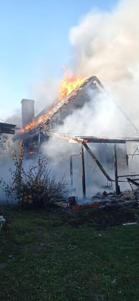 Płonący dach drewnianego domu otoczony gęstym dymem, w tle błękitne niebo.