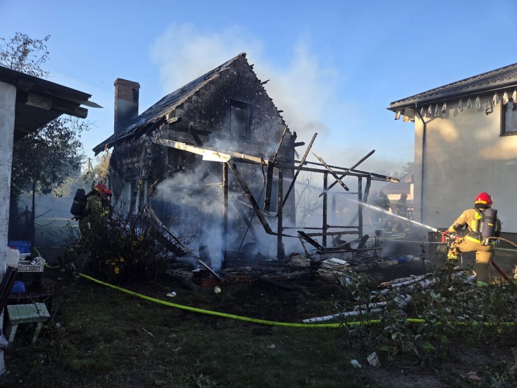 Strażacy gaszą pożar zniszczonego domu, z którego unosi się dym, w otoczeniu roślinności.