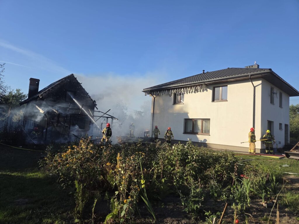 Strażacy gaszą pożar zniszczonego budynku mieszkalnego w otoczeniu ogrodu, w tle widoczny drugi dom.