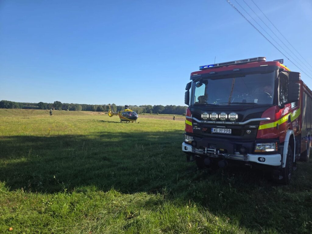 Wielka, czerwona ciężarówka strażacka stoi na zielonym polu, obok lądującego helikoptera ratunkowego.