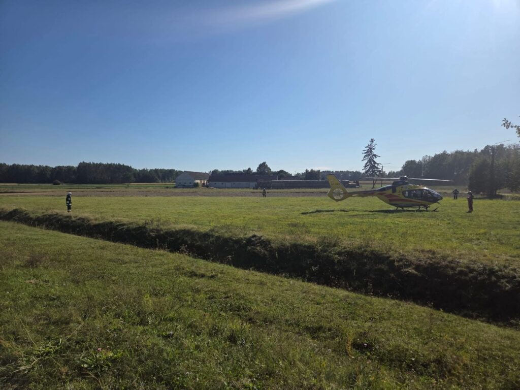 Helikopter ratunkowy ląduje na zielonym polu, obok grupy osób w mundurach i budynku w tle.