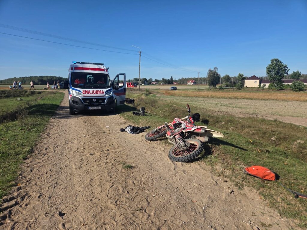 Ambulans z otwartymi drzwiami stoi na piaszczystej drodze obok przewróconego motocykla, a w tle widać grupę ludzi.