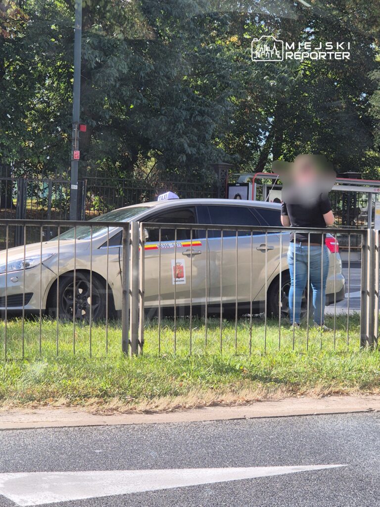 Kobieta w czarnej koszulce stoi obok taksówki zaparkowanej przy chodniku, w tle drzewa i ogrodzenie.