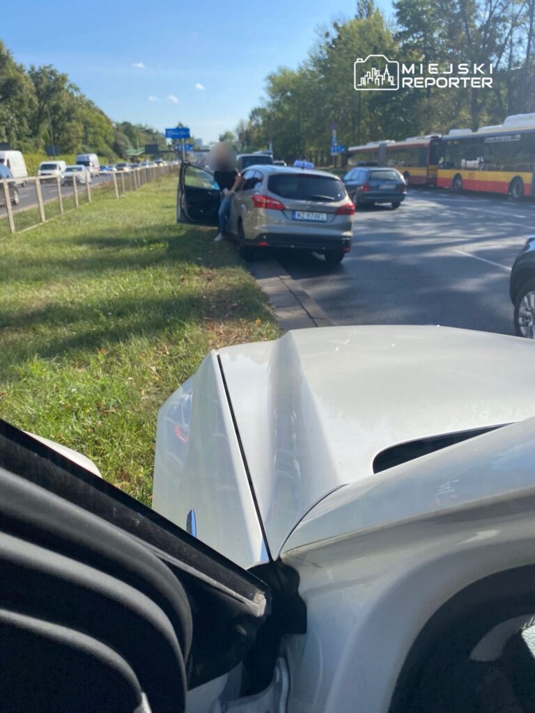 Kobieta w niebieskiej koszuli rozmawia z kierowcą samochodu zaparkowanego na poboczu drogi, obok ruchliwej ulicy.