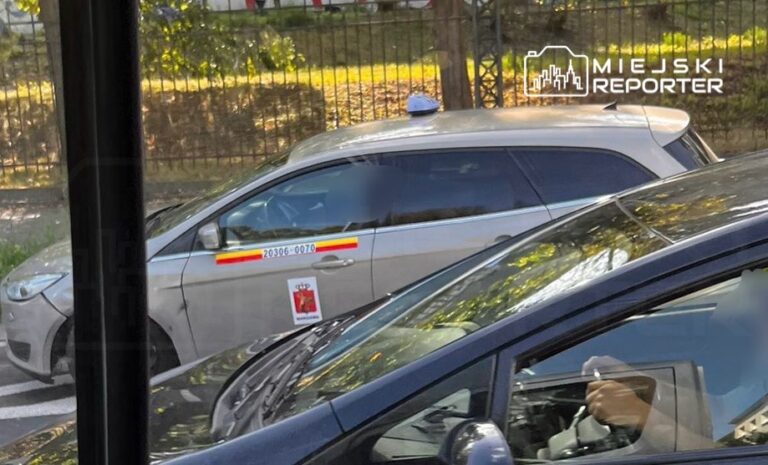 Samochód oznakowany jako radiowóz stoi na ulicy, obok innego pojazdu. Widać osobę siedzącą za kierownicą.