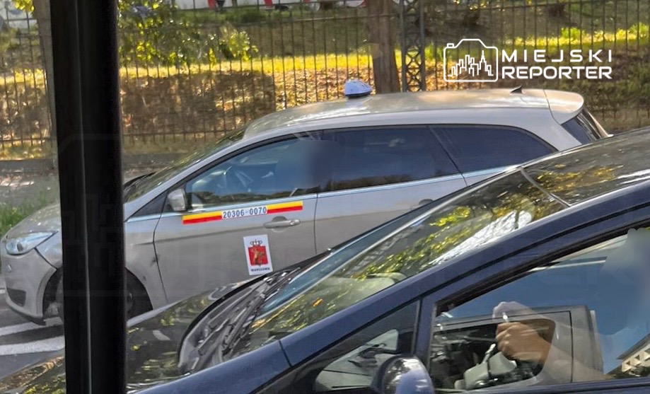 Samochód oznakowany jako radiowóz stoi na ulicy, obok innego pojazdu. Widać osobę siedzącą za kierownicą.