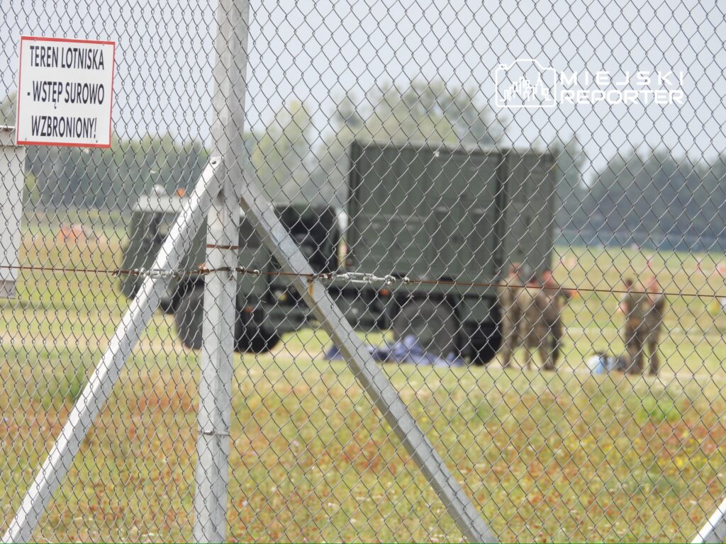 Na terenie lotniska, żołnierze przy ciężarówkach rozmawiają, obok ogrodzenia z siatki. Widoczny znak zakazu wstępu.