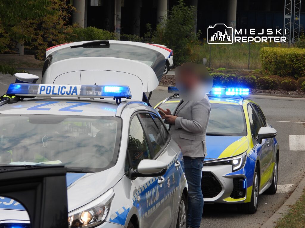 Mężczyzna w szarym garniturze rozmawia przy otwartym bagażniku radiowozu policji na ulicy z włączonymi sygnalizatorami.