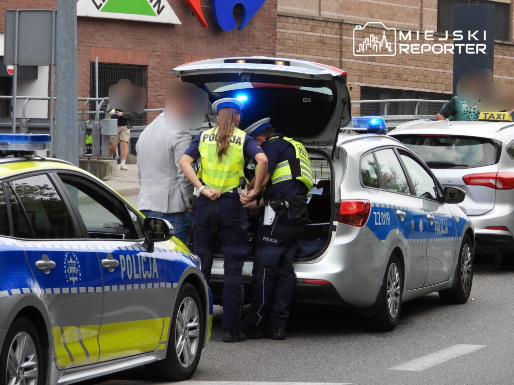 Policjanci zatrzymują osobę w pobliżu radiowozu na miejskiej ulicy, w tle przechodnie i zaparkowane samochody.