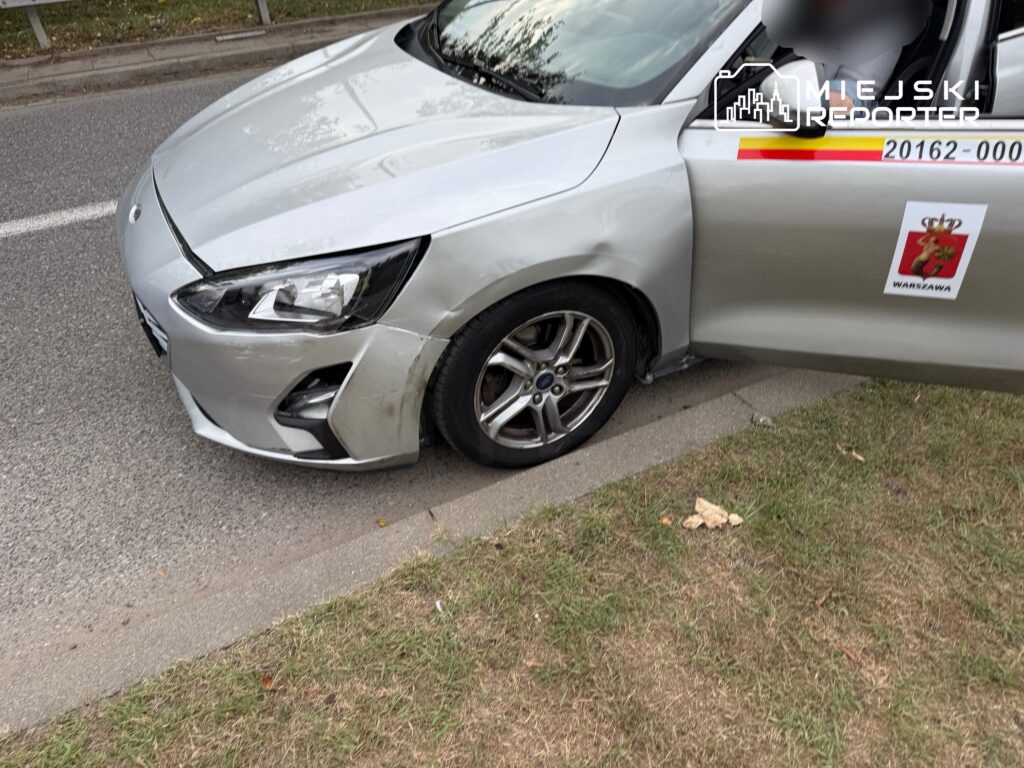 Uszkodzony srebrny samochód zaparkowany na poboczu drogi, z widocznym uszkodzeniem przedniego zderzaka i koła.